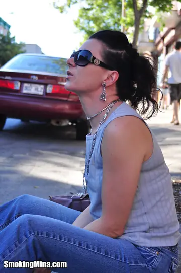 Brunette in Jeans and Glasses Smokes a Cigarette on a Busy Street